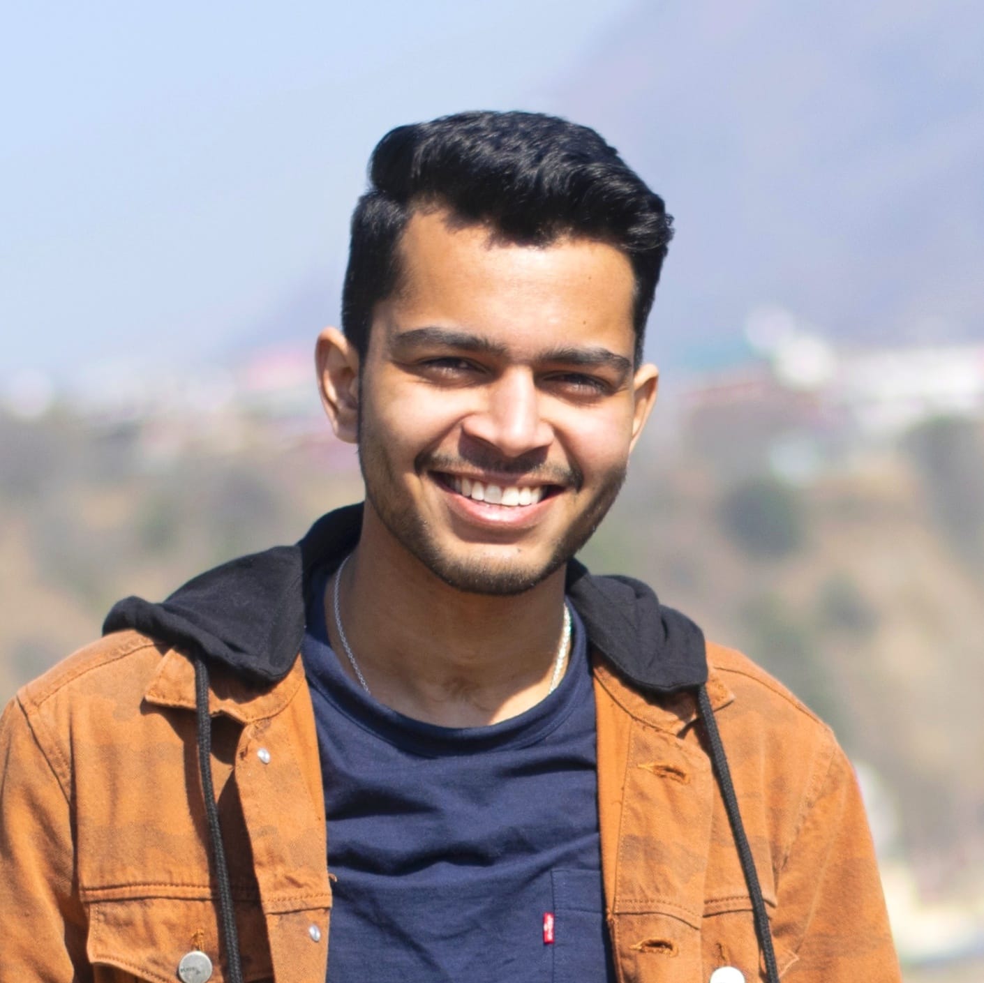 Aditya Raj Singh with short dark hair smiles at the camera outdoors, wearing a brown jacket over a dark blue shirt with a blurred background of nature and distant hills.